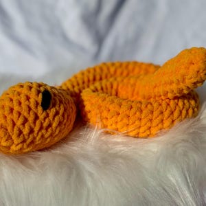 Peut inclure: Un jouet serpent en crochet orange avec des yeux noirs. Le serpent est enroul&eacute; sur une surface blanche et moelleuse.