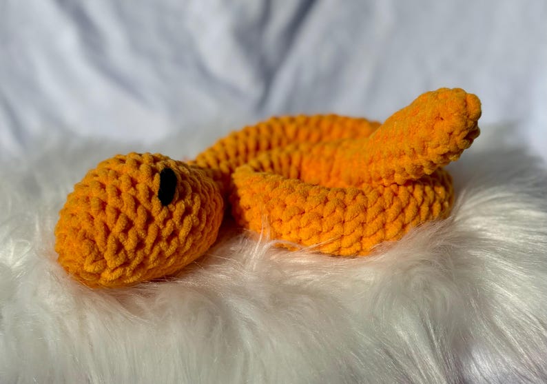 Peut inclure: Un jouet serpent en crochet orange avec des yeux noirs. Le serpent est enroul&eacute; sur une surface blanche et moelleuse.