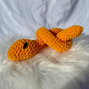 Peut inclure: Un jouet serpent en crochet orange avec des yeux noirs. Le serpent est enroul&eacute; sur une surface blanche et moelleuse.