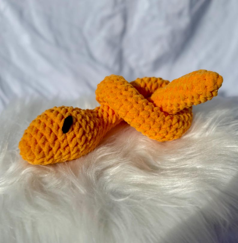 Peut inclure: Un jouet serpent en crochet orange avec des yeux noirs. Le serpent est enroul&eacute; sur une surface blanche et moelleuse.