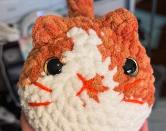 Peut inclure: Un chat en peluche crocheté avec de la fourrure orange et blanche. Le chat a des yeux noirs et un nez et une bouche orange cousus.