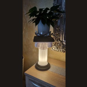 May include: A decorative illuminated column lamp with a white, fluted body and a black base. The lamp has a square black top with a silver lotus flower and a white vase with green leaves. The lamp is on a wooden surface.