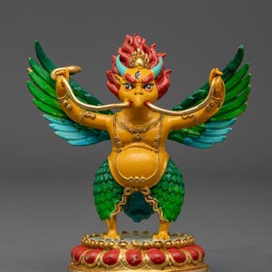 Puede incluir: Una estatua detallada y colorida de una criatura mítica con cuerpo amarillo, alas verdes y azules y cabeza roja y naranja. La figura sostiene una serpiente dorada en cada mano y se alza sobre una base roja y dorada. Es un objeto decorativo.