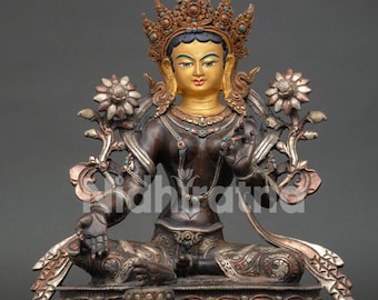 May include: A bronze-toned metal statue of a seated deity with a gold-painted face, wearing an ornate crown and jewellery inlaid with small turquoise stones. The figure is positioned on a lotus base, flanked by two large silver-toned flower stems.