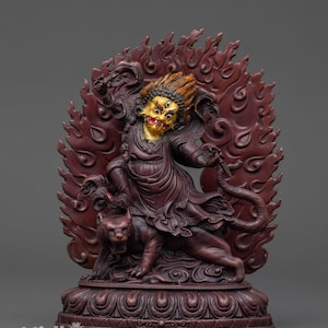 May include: A detailed maroon-colored statue of a deity with a fierce expression, three eyes, and a golden face. The deity is surrounded by a flame-like backdrop and rides a tiger. The statue is on a decorative base. The text "Nidhiratna" is at the bottom.