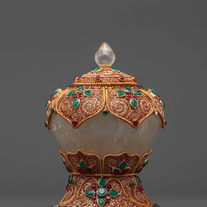 May include: A decorative lidded jar featuring a translucent quartz body encased in intricate gold-toned filigree metalwork. The lid and base are adorned with inlaid green and red teardrop-shaped stones, topped with a clear quartz finial.