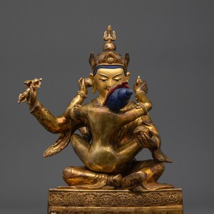 Op de afbeelding: Een gouden beeld van een godheid met meerdere armen, zittend in een meditatieve houding. De figuur draagt een kroon en houdt verschillende voorwerpen vast. Een blauwe schelp wordt bij het gezicht gehouden. Het beeld staat op een decoratieve basis. De tekst "Nidhiratna" staat onderaan.