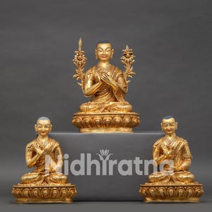 Puede incluir: Tres estatuas doradas de Buda en pose meditativa. La estatua central es más grande y elevada, flanqueada por dos estatuas más pequeñas. Las estatuas están sobre bases ornamentadas, con detalles intrincados. El fondo es gris liso.