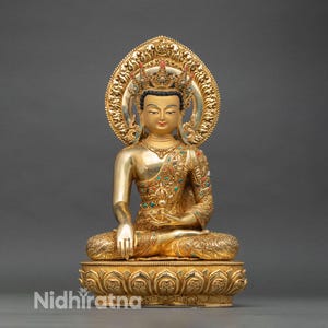 May include: A gold Buddha statue in a seated meditation pose. The statue has detailed features, including a halo, crown, and ornate clothing. The base is a lotus flower design. The statue is set against a grey background.