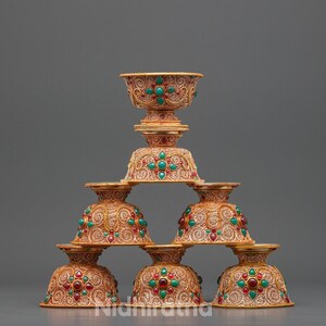 Puede incluir: Una pila piramidal de seis cuencos de metal ornamentados con intrincados patrones de filigrana. Cada cuenco está decorado con piedras redondas rojas y verdes incrustadas en un motivo floral. El metal tiene un acabado dorado cálido.