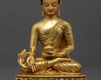 Pode incluir: Uma estátua de Buda de cor dourada na posição sentada de lótus sobre uma base em forma de lótus. A figura tem uma textura de cabelo azul escuro, traços faciais pintados e segura uma pequena tigela na mão esquerda sobre o joelho.