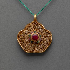 May include: A gold pendant featuring intricate filigree metalwork in a scalloped shape. The center is set with a prominent round red gemstone, surrounded by a cluster of smaller, alternating green and pink circular stones.