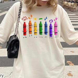 May include: An ivory short-sleeved t-shirt featuring a graphic print of eight colourful crayons with distinct facial expressions and small symbols above each, including a sun, hearts, and clouds, paired with a beige baseball cap and a black shoulder bag.