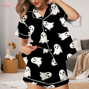 Puede incluir: Un conjunto de pijama negro con un patrón repetitivo de fantasmas blancos con dos ojos negros y una boca ovalada. La camisa de manga corta abotonada tiene un cuello con muescas y ribetes blancos, junto a unos pantalones cortos a juego.