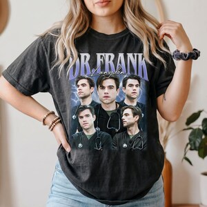May include: A charcoal grey short-sleeve t-shirt featuring a graphic print of a man in medical scrubs with a stethoscope. The text "DR. FRANK" appears in large purple letters above the image, with "Langdon" written in cursive script below.