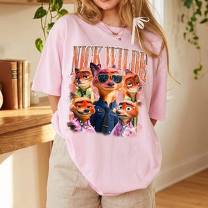 May include: A light pink t-shirt featuring a graphic print of the character Nick Wilde from Zootopia in various poses and expressions. The text NICK WILDE is printed in a large, bold, orange-toned font above the character illustrations.