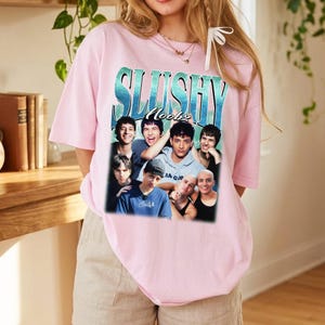May include: A light pink short-sleeve t-shirt featuring a graphic print of six individuals. The text SLUSHY Noobz is printed in a blue gradient font above the group. The design includes a collage of portraits of young people in various poses.