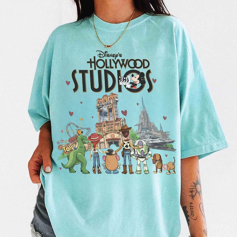 May include: Light blue t-shirt featuring the text "Disney's Hollywood Studios" above a graphic of Disney characters and park attractions. The graphic includes Mickey Mouse, Toy Story characters, and the Tower of Terror.