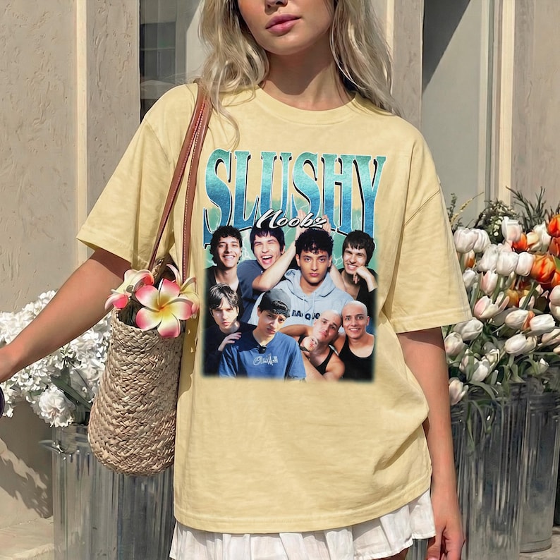 May include: A pale yellow short-sleeve t-shirt featuring a graphic print with the text SLUSHY and Noobz above a collage of seven individuals. The shirt has a crew neckline and a relaxed fit, worn with a woven straw bag featuring pink flowers.