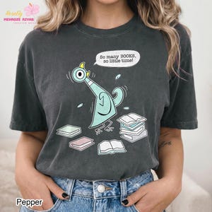 May include: A charcoal gray short-sleeve t-shirt featuring a graphic of a light blue pigeon surrounded by several books. A speech bubble above the pigeon contains the text, "So many BOOKS, so little time!" The shirt has a crew neckline and a relaxed fit.
