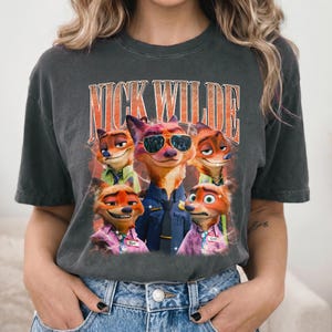 May include: A charcoal gray short-sleeve t-shirt featuring a graphic print of the character Nick Wilde from Zootopia in various poses. The text NICK WILDE is printed in large orange letters above the characters, who are wearing shirts and ties.