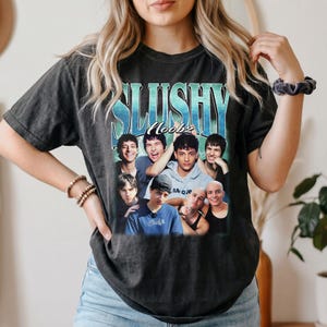 May include: A charcoal grey short-sleeve t-shirt featuring a graphic print with the text SLUSHY Noobz in blue and white. The design includes a collage of six individuals in various poses and expressions, set against a light blue lightning background.