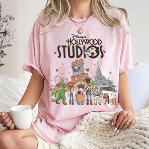May include: A light pink t-shirt featuring the text "Disney's Hollywood Studios" with Mickey Mouse and various Toy Story characters. The graphic includes the Tower of Terror and other park attractions. The shirt is a comfortable, casual style.