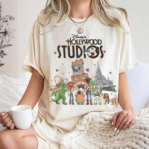 May include: Cream-colored t-shirt featuring the text "Disney's Hollywood Studios" with Mickey Mouse and various cartoon characters. The graphic includes images of the Tower of Terror, a roller coaster, and other park attractions. Small red hearts are scattered around the design.
