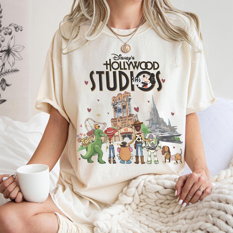 May include: Cream-colored t-shirt featuring the text "Disney's Hollywood Studios" with Mickey Mouse and various cartoon characters. The graphic includes images of the Tower of Terror, a roller coaster, and other park attractions. Small red hearts are scattered around the design.