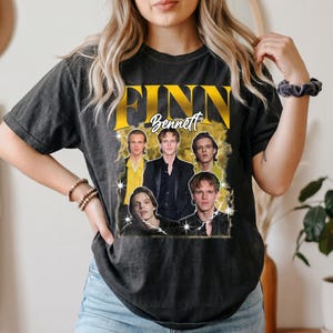 Könnte beinhalten: Ein anthrazitfarbenes Kurzarm-T-Shirt mit einem großen Grafikdruck, der den Text "FINN Bennett" in fetten gelben Buchstaben zeigt. Das Design enthält vier verschiedene fotografische Porträts derselben Person in einer Collage.