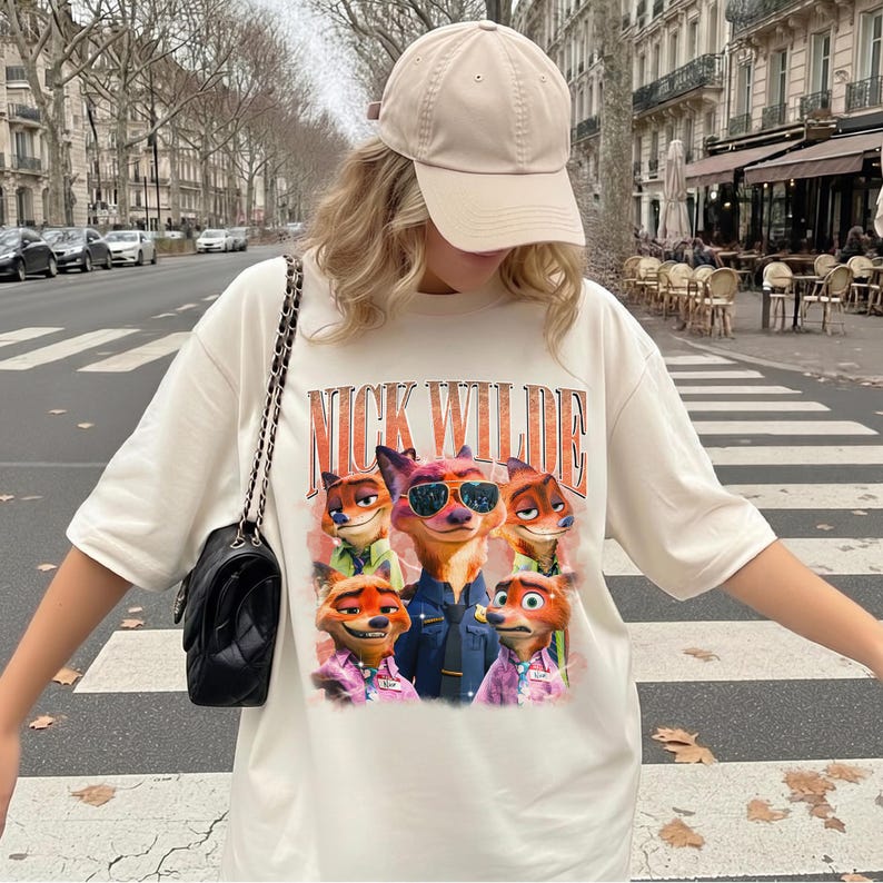 May include: A cream-colored short-sleeve t-shirt featuring a graphic print of the character Nick Wilde from Zootopia in various poses and outfits, with the text NICK WILDE printed in large orange letters above the character illustrations.