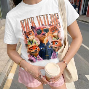 May include: A white short-sleeved t-shirt featuring a graphic print of the character Nick Wilde from Zootopia in various poses and expressions. The text NICK WILDE is printed in large, bold, orange-outlined letters above the character illustrations.