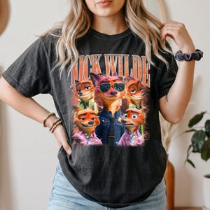 May include: A charcoal gray short-sleeve t-shirt featuring a graphic print of the character Nick Wilde from Zootopia in various poses and outfits. The text NICK WILDE is printed in large orange letters above the character illustrations.
