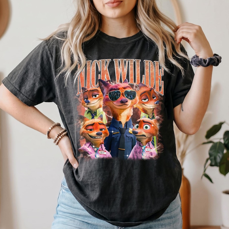 May include: A charcoal gray short-sleeve t-shirt featuring a graphic print of the character Nick Wilde from Zootopia in various poses and outfits. The text NICK WILDE is printed in large orange letters above the character illustrations.