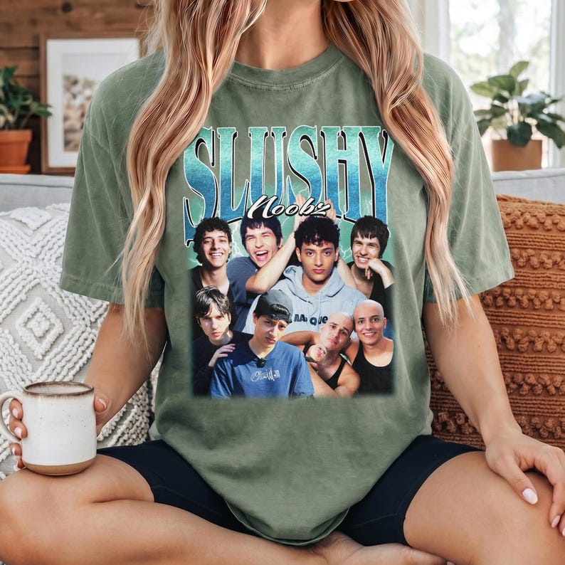 May include: A sage green short-sleeve t-shirt featuring a graphic print with the text SLUSHY and Noobz above a collage of seven individuals. The design includes a blue gradient background behind the text and a group photograph of people.
