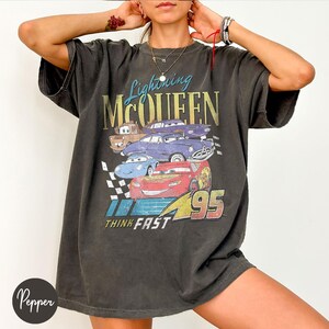 May include: Dark gray Lightning McQueen graphic t-shirt featuring various cartoon cars. The design includes the text "Lightning McQueen," "Think Fast," and the number "95." The shirt has a vintage, worn look.