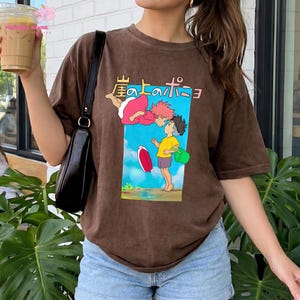 May include: A brown short-sleeve t-shirt featuring a graphic print of two animated characters, one with pink hair and one with brown hair, against a blue sky background. The Japanese text 崖の上のポニョ is printed above the characters.
