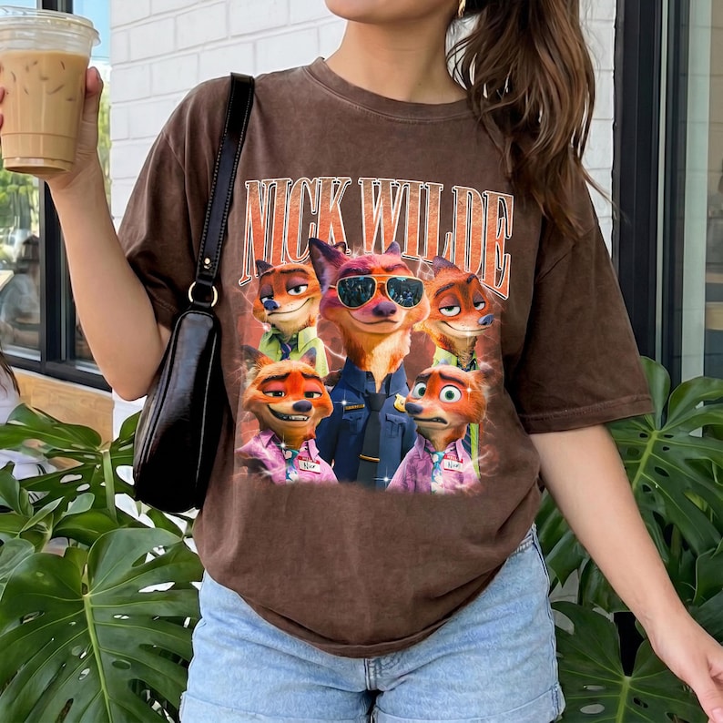 May include: A brown short-sleeve t-shirt featuring a graphic print of the character Nick Wilde from Zootopia in various poses and expressions. The text NICK WILDE is printed in a large, bold, orange-outlined font above the character illustrations.