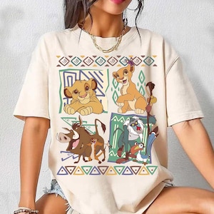 May include: Cream-colored t-shirt featuring a colorful graphic of characters from The Lion King. The design includes Simba, Nala, Timon, Pumbaa, Rafiki, and Zazu, set against geometric patterns. The shirt has short sleeves and a crew neck.