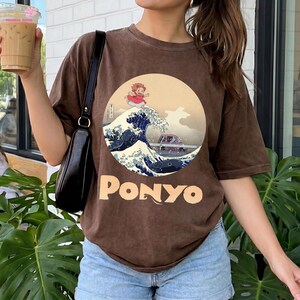 May include: A brown short-sleeve t-shirt featuring a circular graphic of a character running on a large ocean wave with a small purple car nearby. The word PONYO is printed in large beige letters below the circular illustration on the front.
