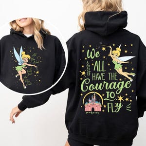 Op de afbeelding: Zwarte hoodie met een cartoon fee ontwerp en de tekst "All You Need Is Faith, Trust, And Pixie Dust". De achterkant van de hoodie heeft de tekst "We All Have The Courage To Fly" met een kasteelafbeelding.
