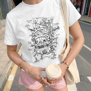 May include: A white short-sleeved t-shirt featuring a detailed black line art illustration of a complex mechanical castle structure. The graphic includes handwritten labels such as Discharge pipe, Helix direction, Wind gauge, Dwelling house, and Secondary propeller.