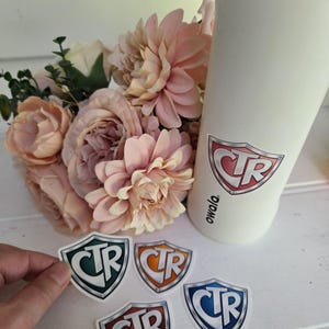 Op de afbeelding: Een witte geïsoleerde waterfles met een CTR-logosticker, naast een boeket roze bloemen en verschillende CTR-logostickers in verschillende kleuren. De stickers zijn schildvormig met de letters CTR.