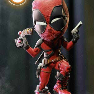 May include: A red and black Deadpool figurine, holding a gun and a sign that says "Click!". The figure is standing on a detailed base with a metallic and textured design. The overall theme is comic book characters.
