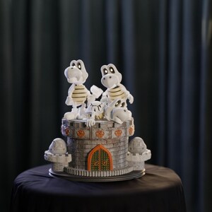 May include: A detailed cake featuring Dry Bones characters from the Super Mario franchise. The cake is designed as a castle with a gray brick exterior, orange accents, and a green and orange door. The Dry Bones figures are white with tan stripes.
