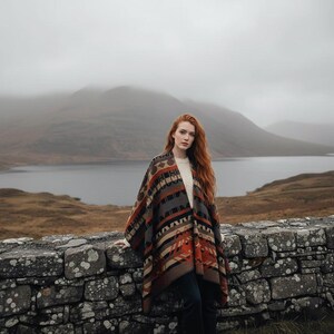 Op de afbeelding: Een patroonponcho in bruin-, oranje- en grijstinten. De poncho heeft een geometrisch ontwerp en wordt over een witte trui gedragen. De achtergrond omvat een stenen muur, een meer en bergen onder een bewolkte hemel.