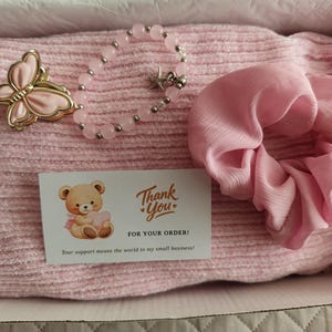 May include: A pink ribbed fabric item, a pink butterfly-shaped hair clip, a beaded bracelet with a small starfish charm, a pink fabric scrunchie, and a thank you card featuring a teddy bear illustration and the text Thank You FOR YOUR ORDER.