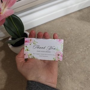 May include: A rectangular white thank you card featuring a floral border with pink blossoms and green leaves. The text reads: Thank You FOR YOUR ORDER! Your support means the world to my small business! The card is held against a neutral background.