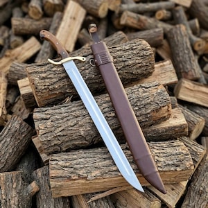 May include: A steel sword with a polished blade, a brass crossguard, and a wooden handle secured with metal rivets. The sword is accompanied by a brown leather scabbard featuring stitched detailing, a metal ring, and a top strap with a metal button.