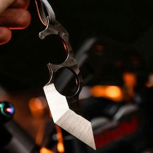 May include: A dark metal karambit knife with a textured finish and a lanyard. The blade has a sharp, angled edge. The knife is held against a blurred background with hints of orange and black.